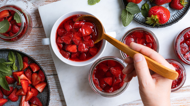 Top view of a wooden counter with a marble board and several jars and a bowl of strawberry compote and some fresh strawberries on the side