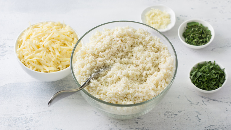 Cauliflower rice in clear bowl surrounded by small bowls of herbs and shredded cheese