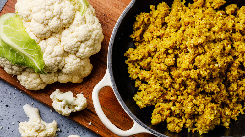 Yellow seasoned cauliflower rice in cast iron pan next to head of whole cauliflower