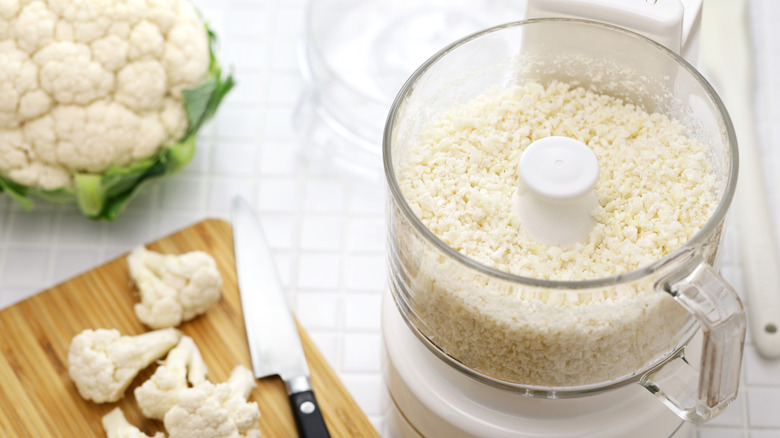 Cauliflower rice in food processor next to fresh cauliflower heads