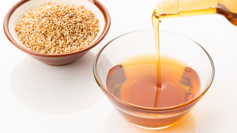 Sesame oil being poured into small glass bowl next to ceramic bowl of sesame seeds