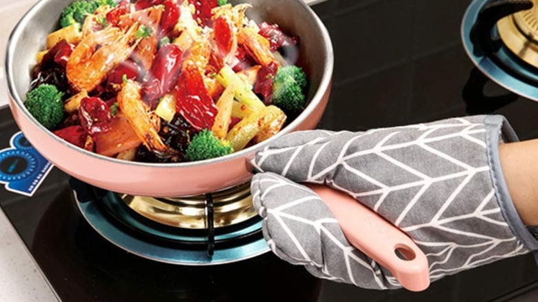 Hand wearing oven mitts holding a pan of food over stove