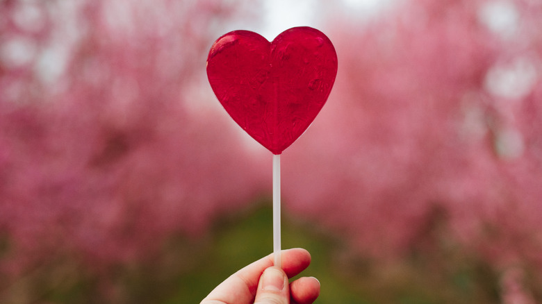 Hand holding a heart-shaped lollipop against a blurred pink background