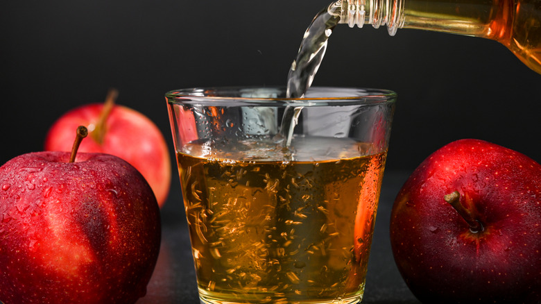 Apple cider vinegar being poured into a glass with three apples in the background
