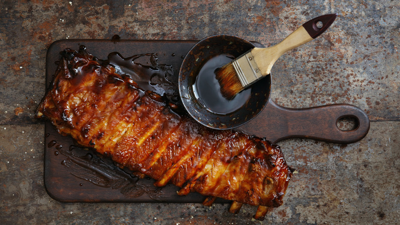 Grilled spicy glazed pork ribs on a wooden board next to a bowl of glaze with a brush