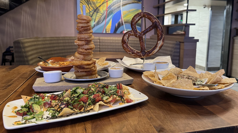 A tower of onion rings, a large pretzel, and nachos are displayed on white plates on a table.