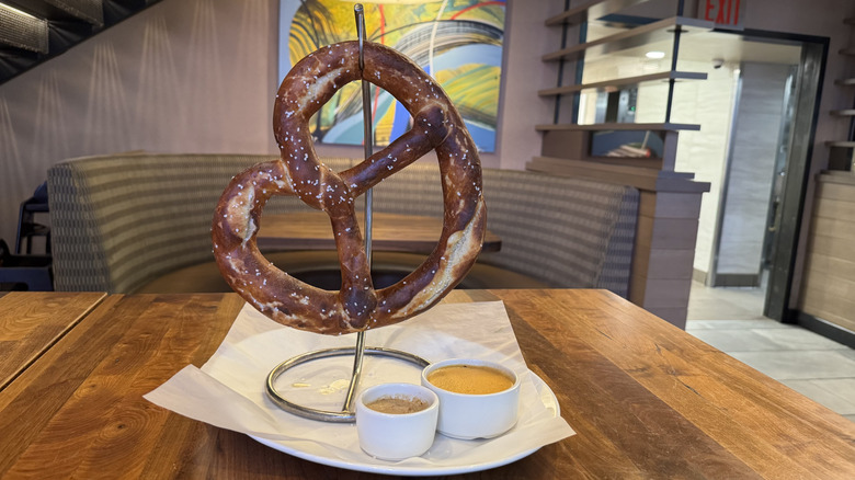 An extra large pretzel is hanging above a white plate with two sauces on a wood table.