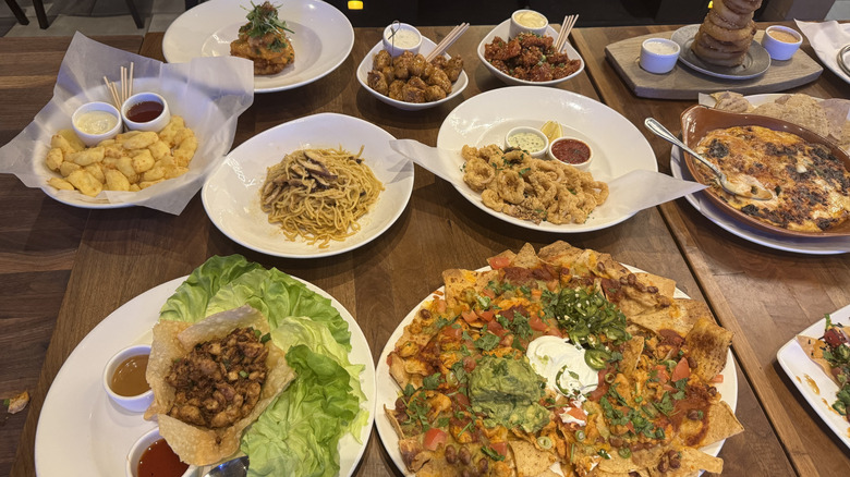 A wooden table is full of appetizers on white plates: fried cheese, nachos, lettuce wraps, pasta, and wings.