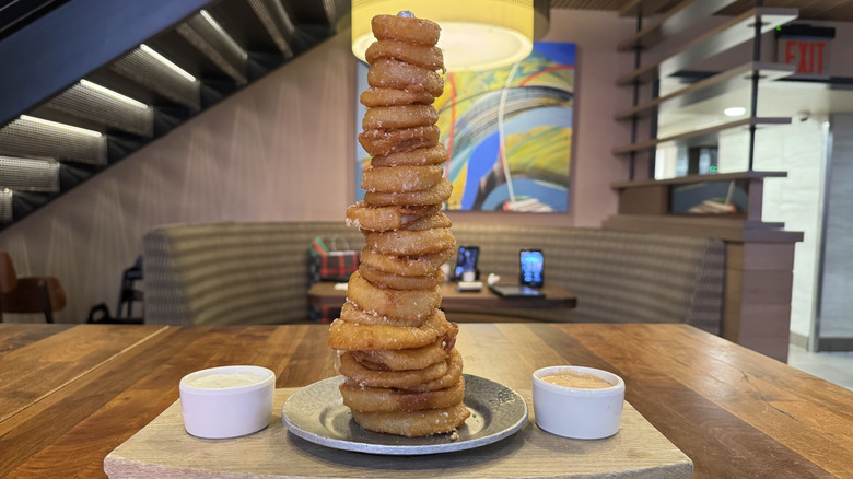 A tower of golden onion rings sits on a wooden slab with two sauces.