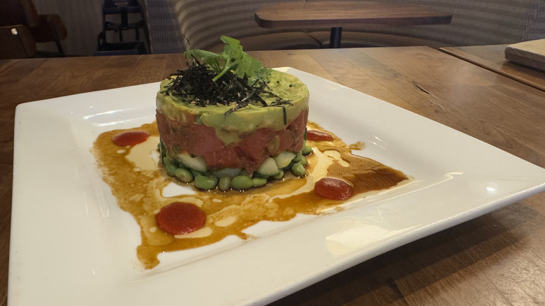 A square white plate holds a tower of raw tuna with avocado and edamame on a wood table.