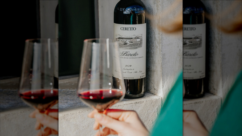 a hand holding red wine glass with a Barolo red wine bottle in the back