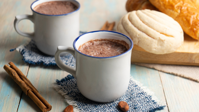 Two cups of hot chocolate next to bread and a cinnamon stick