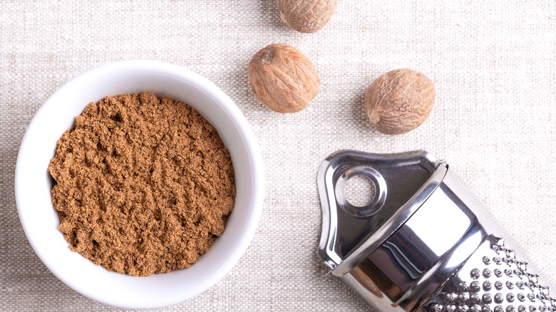 A bowl of ground nutmeg next to whole nutmeg and grater