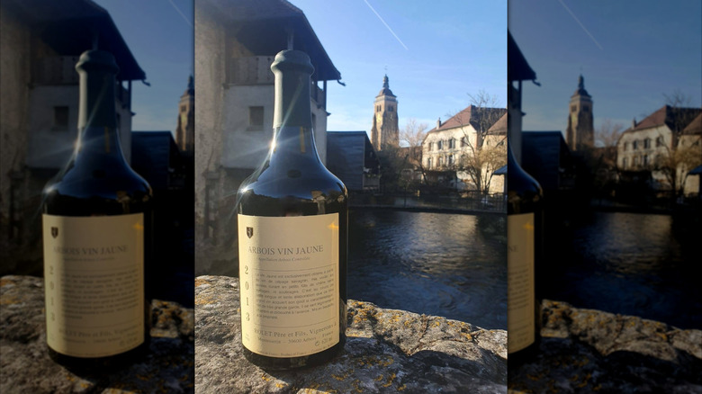 A bottle of Domaine Rolet Arbois vin jaune set on a stone bridge with water in the background