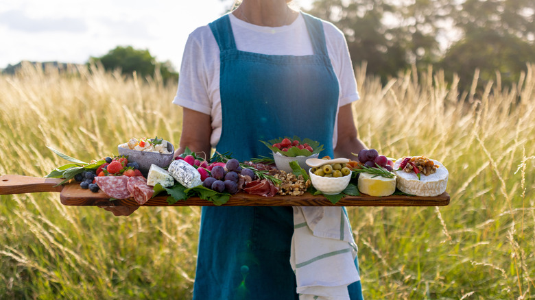 Person holding a charcuterie board with cheese in front of field