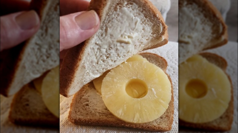 Hand lifting slice of bread on sandwich to expose pineapple rings and mayo