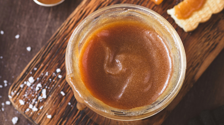 Creamy, smooth homemade salted caramel inside a glass jar with salt sprinkled on the side, all set on a wood background