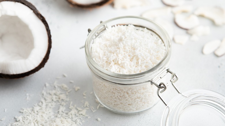 A glass jar with an airtight lid filled with coconut flakes, placed on a white background surrounded by raw coconut halves and sprinkled coconut flakes