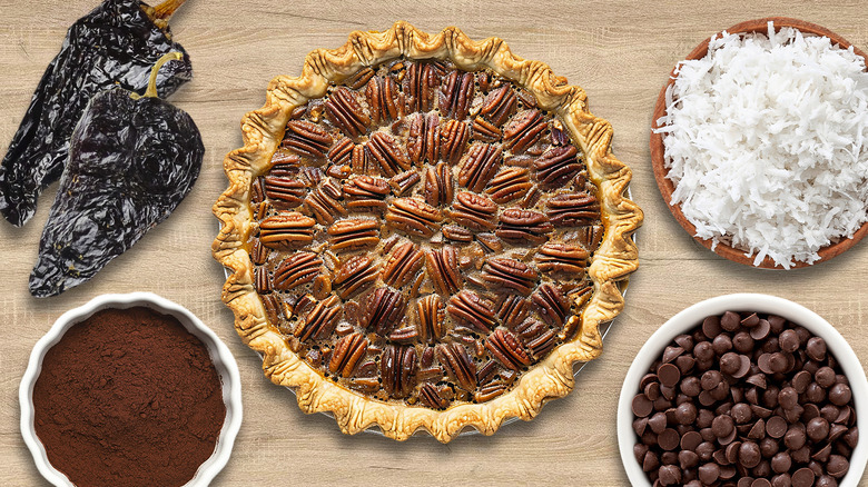 Classic pecan pie in the center surrounded by desiccated coconut, chocolate drops, espresso powder, and ancho chili peppers, all assembled on a wood background