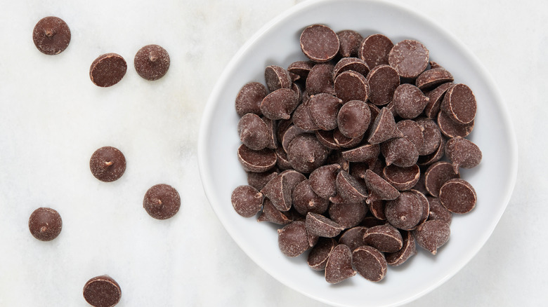 Chocolate chips in a white bowl, surrounded by chocolate chips spread on a white background