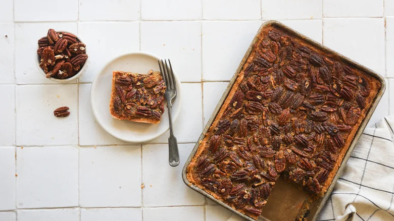 Classic pecan pie made in the form of a slab pie with one piece taken out on a plate placed next to a bowl of pecans, all set on a white background