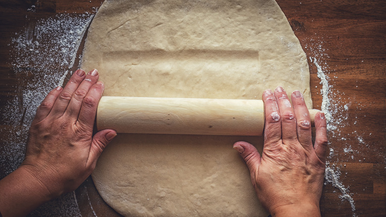 Top view of female hands rolling pie crust with a rolling pin on a dark brown background