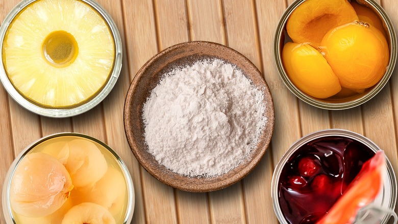 Boxed cake mix in a wooden bowl surrounded by canned pineapple, peaches, cherries, and pears on a wooden table