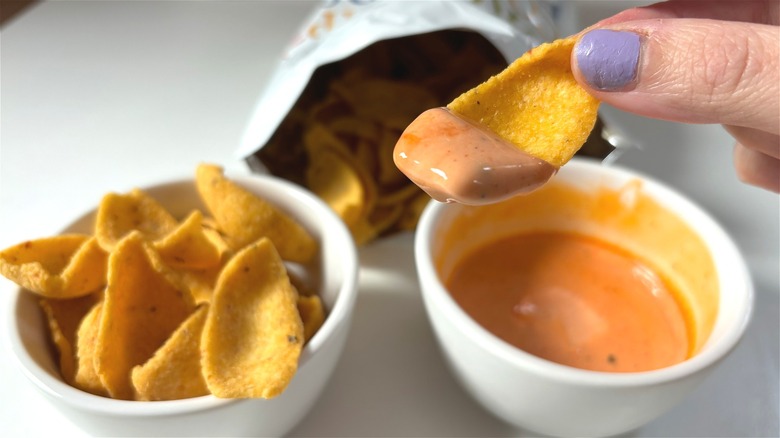 holding trader joes corn chip dipper with sauce, cup of chips, sauce, and bag of chips in background