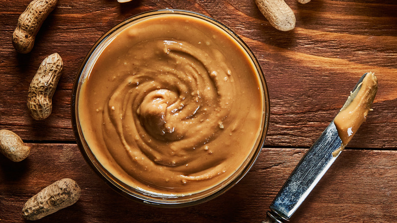 Bowl of nut butter surrounded by peanuts on wooden surface