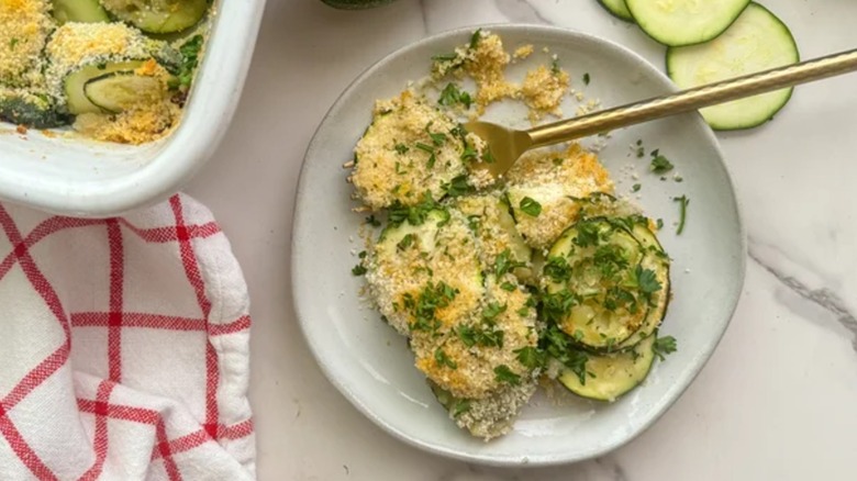 Zucchini gratin on a white plate with a golden fork on a white counter with sliced zucchinis
