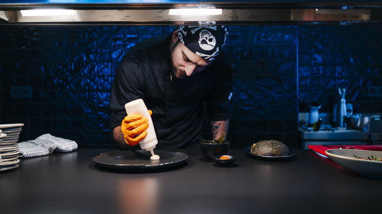 Chef in a modern kitchen plating a dish under dramatic lighting. The chef, wearing a black uniform, orange gloves, and a bandana, uses a squeeze bottle to carefully add sauce to a dark plate. The background features a sleek, tiled wall and various kitchen tools and ingredients, creating a focused and artistic atmosphere.