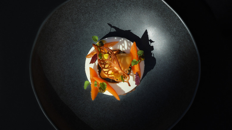 Artistic dessert presented in a deep black bowl, featuring a circular white base topped with a caramelized garnish, delicate orange segments, and vibrant microgreens. The plating is illuminated dramatically, with shadows emphasizing the textures and colors against the dark, minimalist background, creating a striking visual contrast.