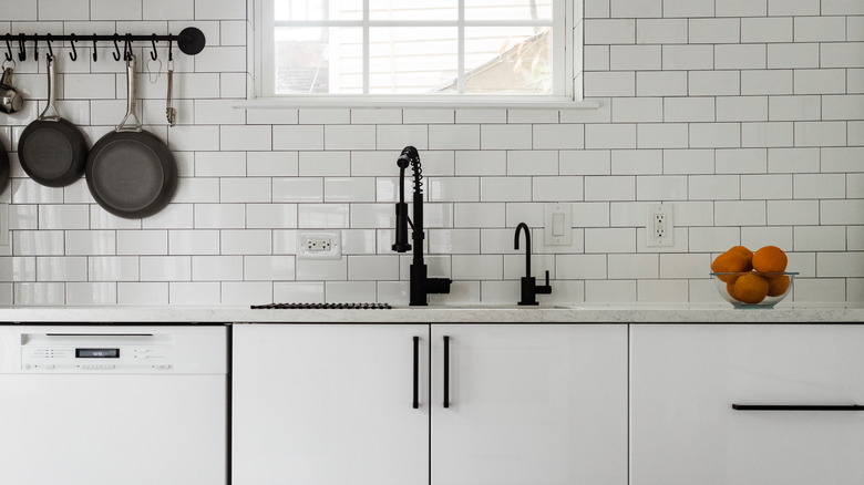 Front view of a white kitchen counter, sink, hanging pot rack, a glass bowl with oranges with white subway tile backsplash