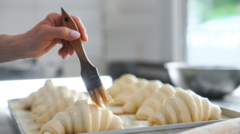 Person using pastry brush on tray of croissants