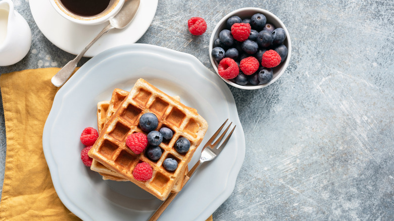 Belgian waffles are topped with berries with a cup of coffee on the side.