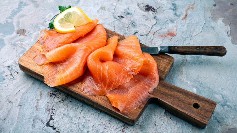 Smoked salmon sits in slices with parsley and lemon on a cutting board.