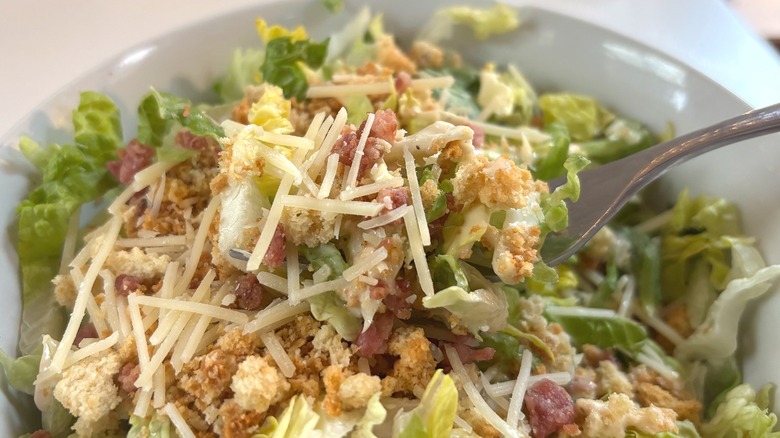 Fork holding bacon Caesar salad bite above white bowl of Caesar salad