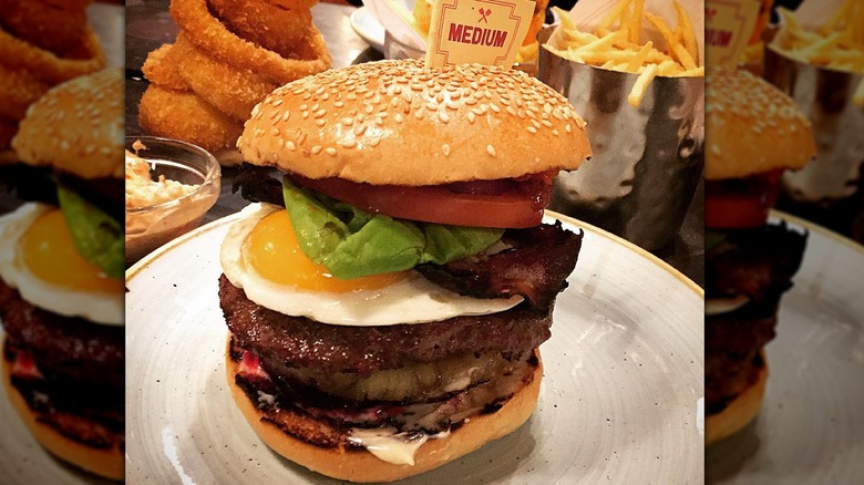 Tall burger on white plate near onion rings and fries