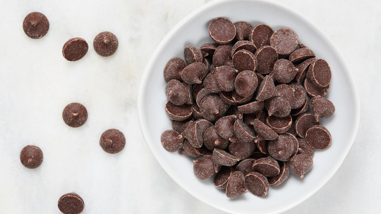 Dark chocolate chips in a bowl surrounded by several chocolate chips spread on the white background