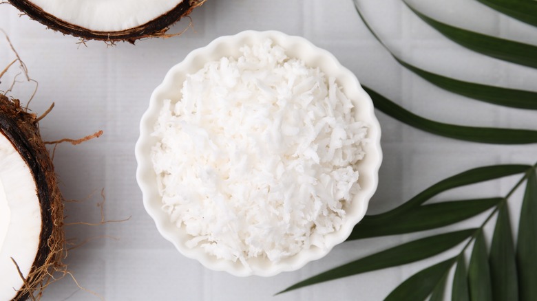 Dried coconut flakes in a white bowl with a white background, surrounded by raw coconut halves