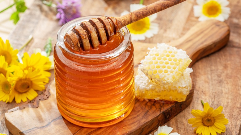 Natural organic honey in a jar holding a wooden honey dipper, surrounded by honeycomb and flowers