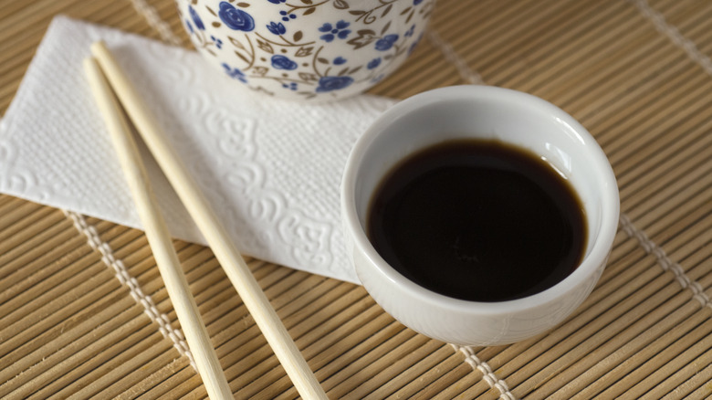 White ceramic bowl filled with dark brown soy sauce placed on a bamboo mat next to chopsticks with a white tissue and a floral bowl in the back