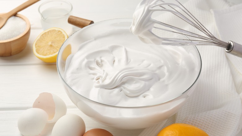 Fresh whipped cream in a glass bowl with a whisk and a white background, surrounded by sugar, a lemon half, and cracked egg shells