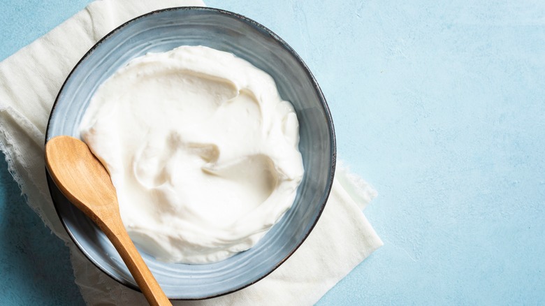 A round bowl filled with firm, white yogurt, accompanied by a wooden spoon, all set on a white towel and a blue background