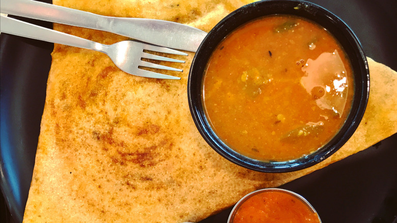 Sambar in a black bowl with dosa and utensils