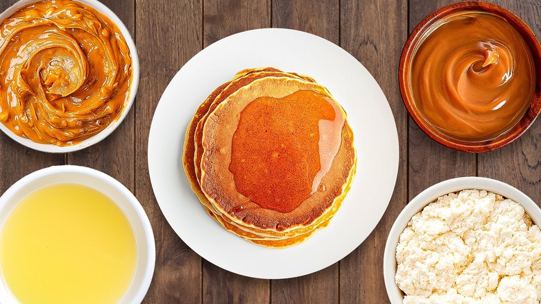 A stack of pancakes with maple syrup is surrounded by bowls with other toppings.