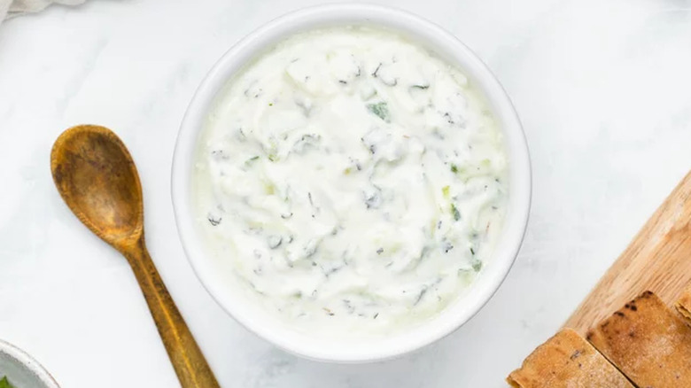 Bowl of tzatziki on marble next to wooden spoon and sliced bread