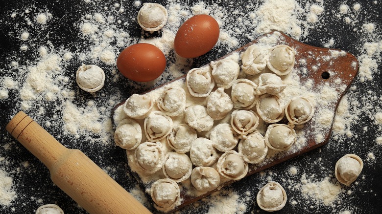 Uncooked pelmeni on cutting board with rolling pin, eggs, and flour on the side