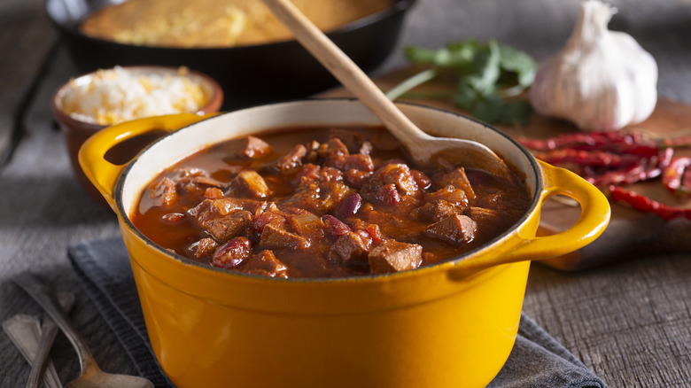 Yellow pot of chili con carne with wooden spoon, and chilis and garlic in the background