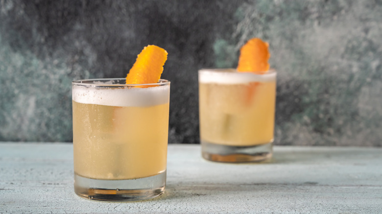 Two whiskey sours in tumbler glasses with egg white froth and orange peel garnishes